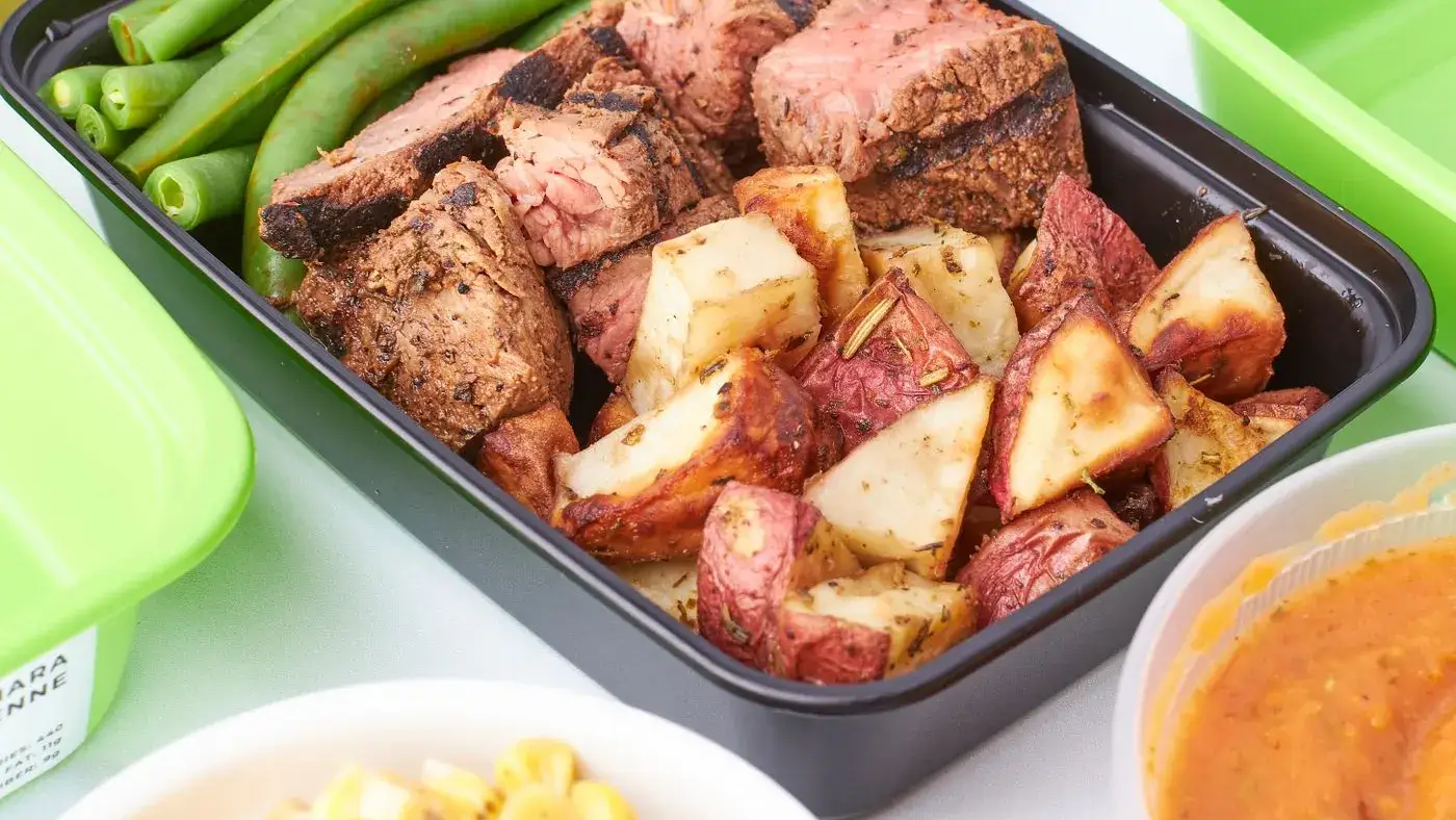 Recipiente de almuerzo con carne asada, papas al horno y habichuelas, junto a un tazón de maíz