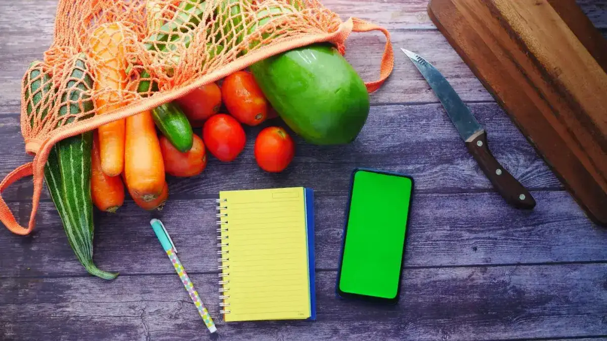 Bolsa de red con zanahorias, tomates, pepino y papaya sobre una mesa de madera, junto a una libreta amarilla, un lápiz y un celular, para anotar las cuentas de las calorías del día