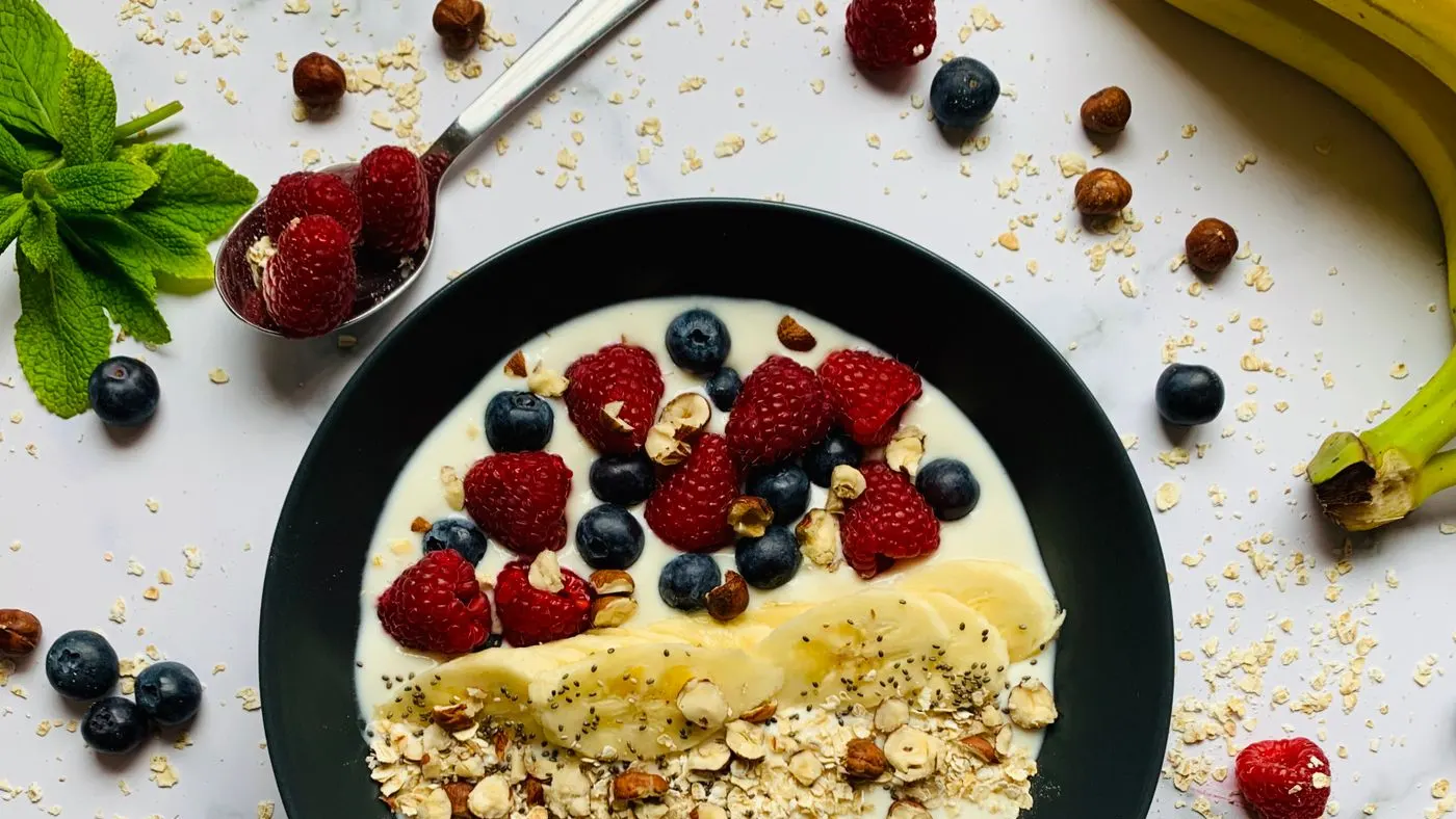 Tazón negro con yogur, banano, frambuesas, arándanos, avena y avellanas sobre una mesa blanca