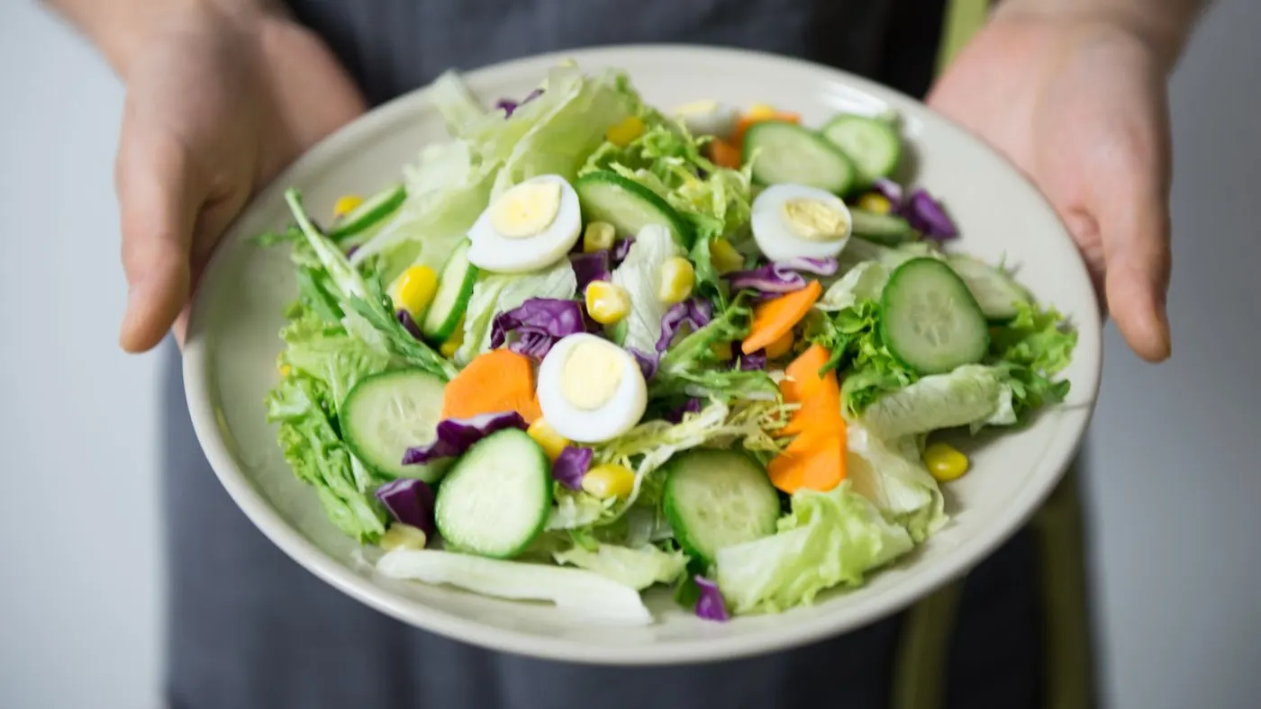 Dos manos sostienen un plato de ensalada con lechuga, pepino, zanahoria, repollo morado, maíz y huevo de codorniz
