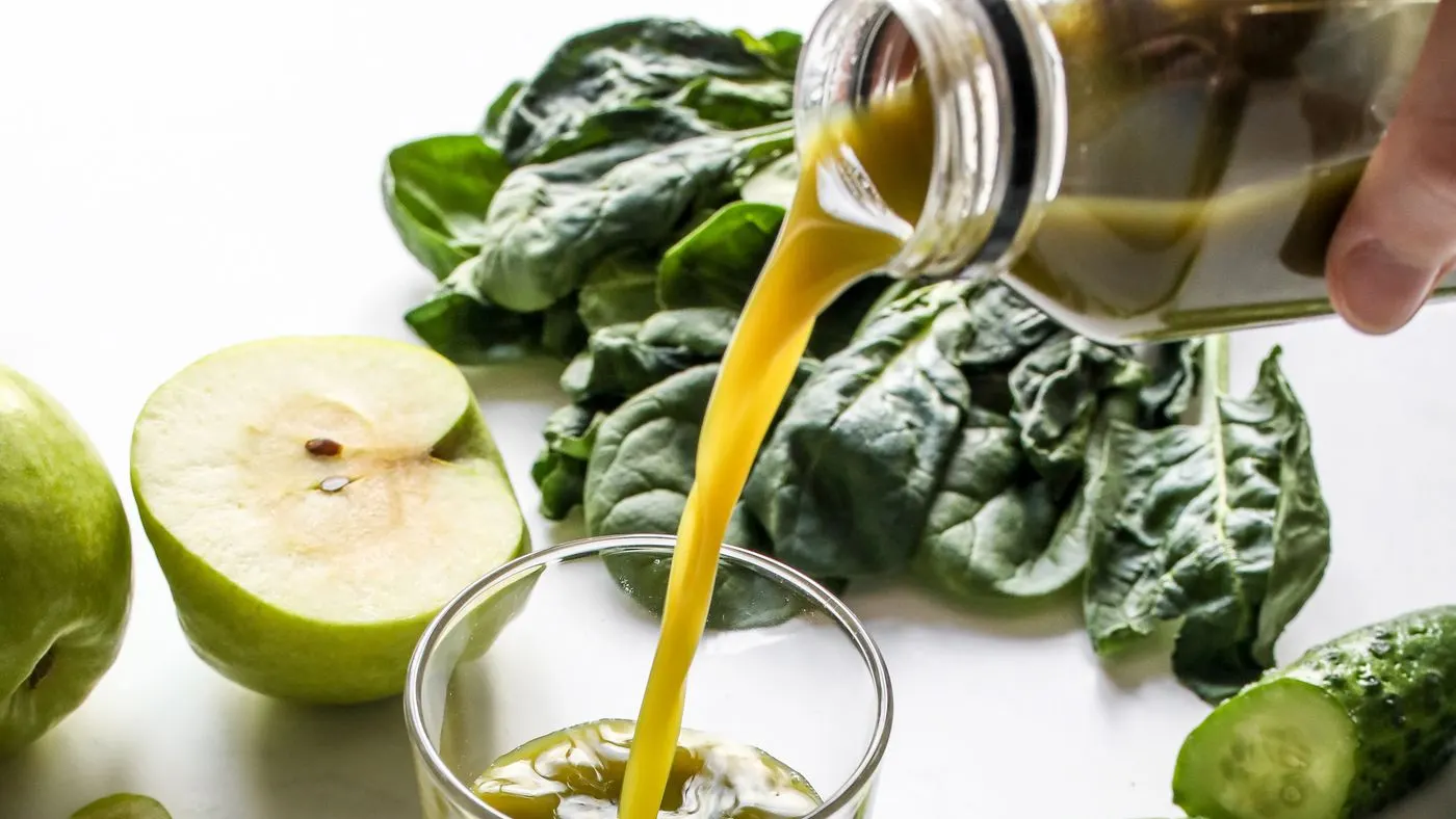 Jugo verde que se sirve desde una botella de vidrio a un vaso, con hojas de espinaca, media manzana verde y un trozo de pepino alrededor