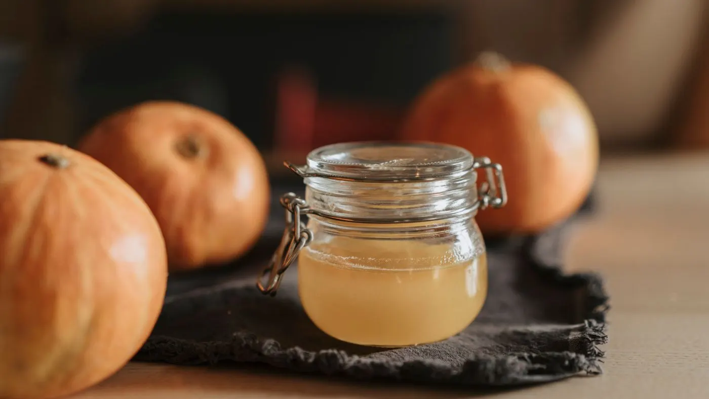 Frasco de vidrio con vinagre de manzana, un líquido dorado, entre calabazas sobre una mesa de madera